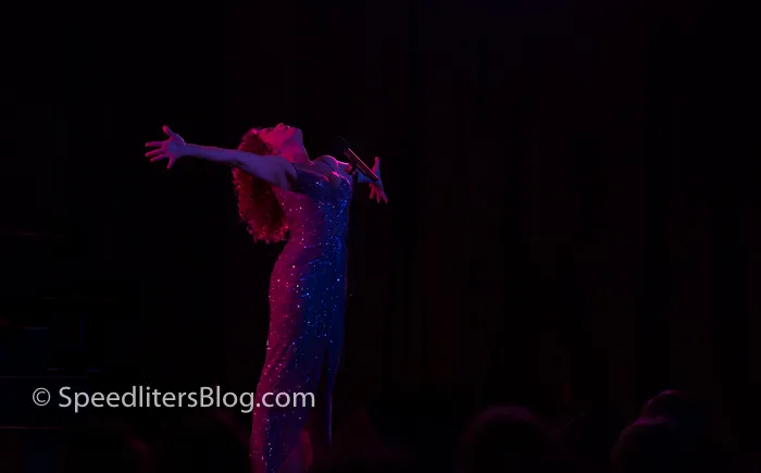 Bernadette Peters in a sequined gown with arms outstretched under red stage light