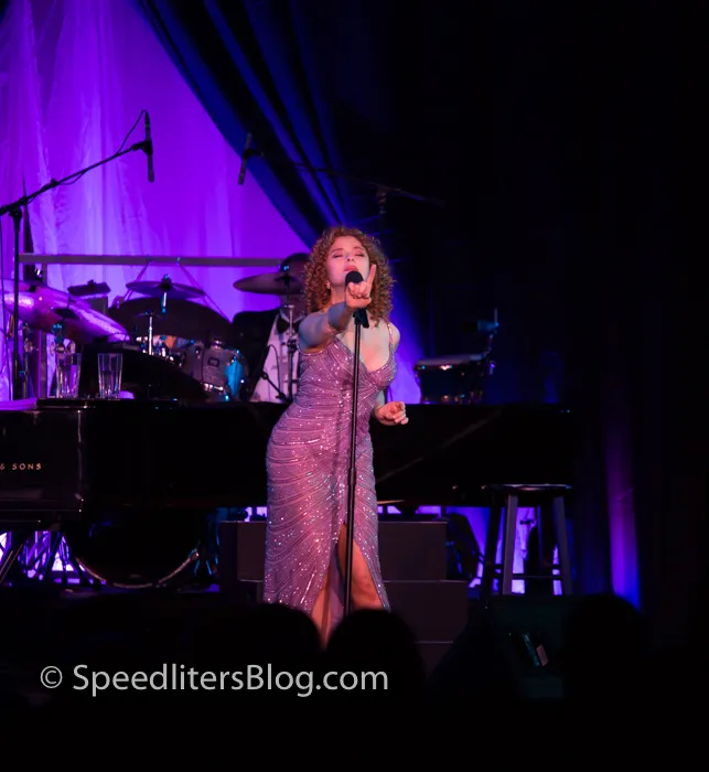Bernadette Peters singing in a sparkling gown in front of a band and purple stage lights