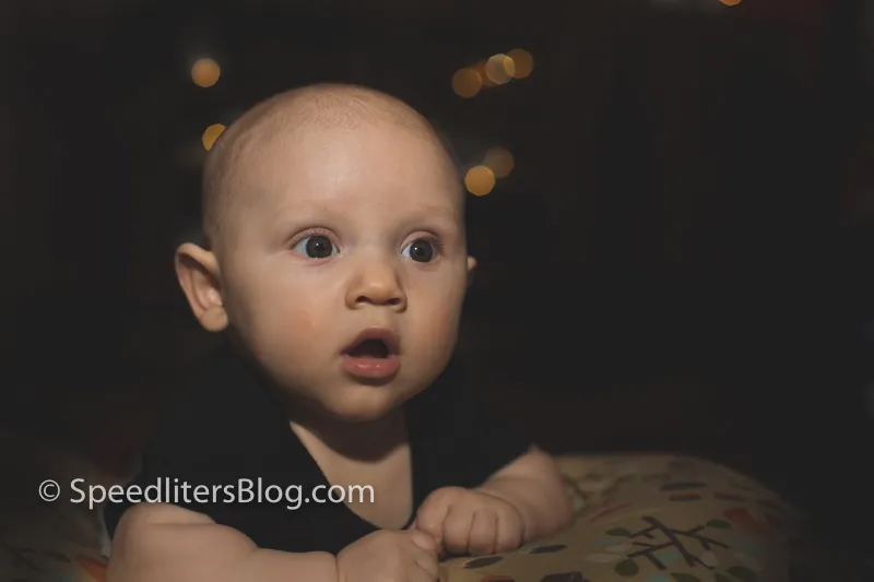 Wide-eyed baby lying on a pillow, lit by bare speedlite with bokeh behind