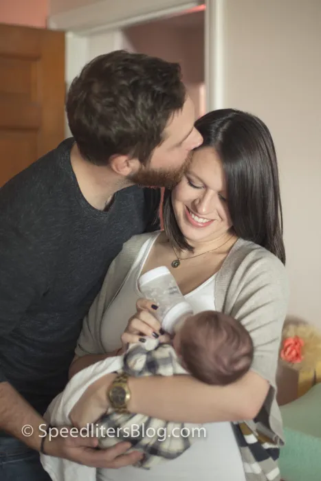 Father kissing mother’s cheek as she bottle-feeds their newborn by a window