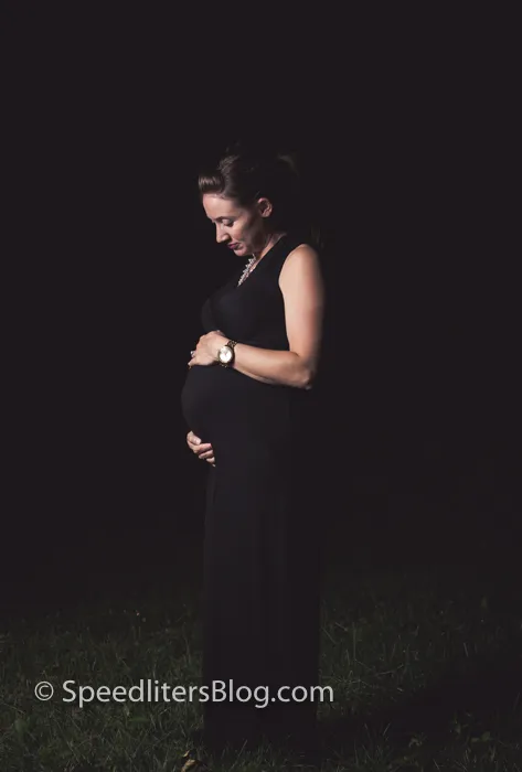 Pregnant woman in a black dress cradling her belly at dusk, lit by one speedlite