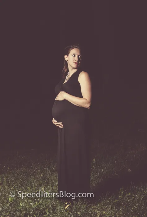 Maternity portrait of a woman in a black dress in a dark field, lit by an umbrella