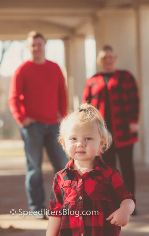 Toddler in a red plaid shirt with her parents blurred behind her