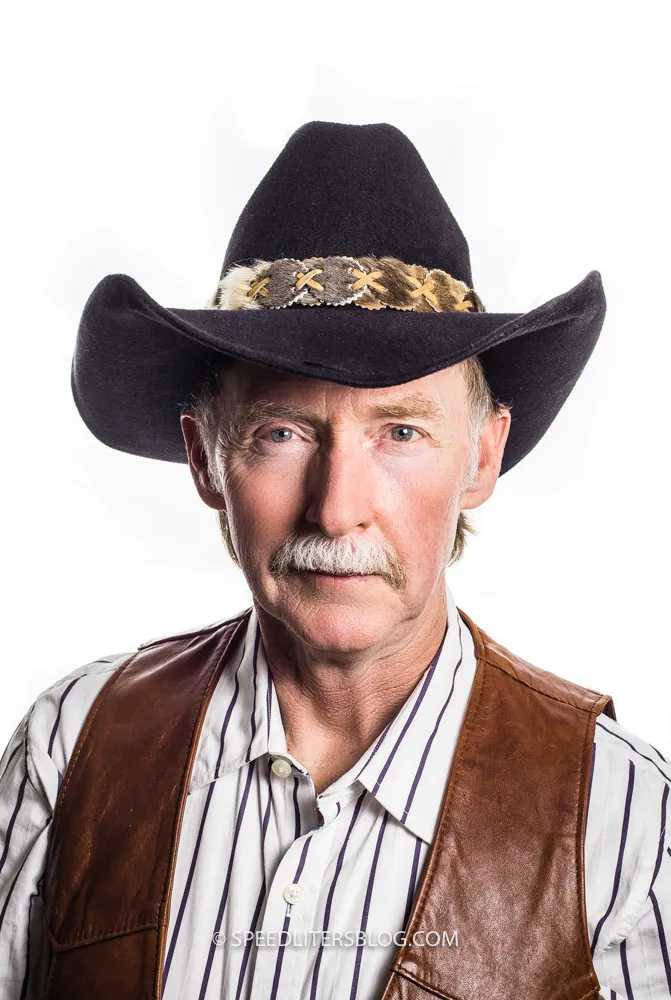 Older man in a black cowboy hat and brown leather vest against a white umbrella background