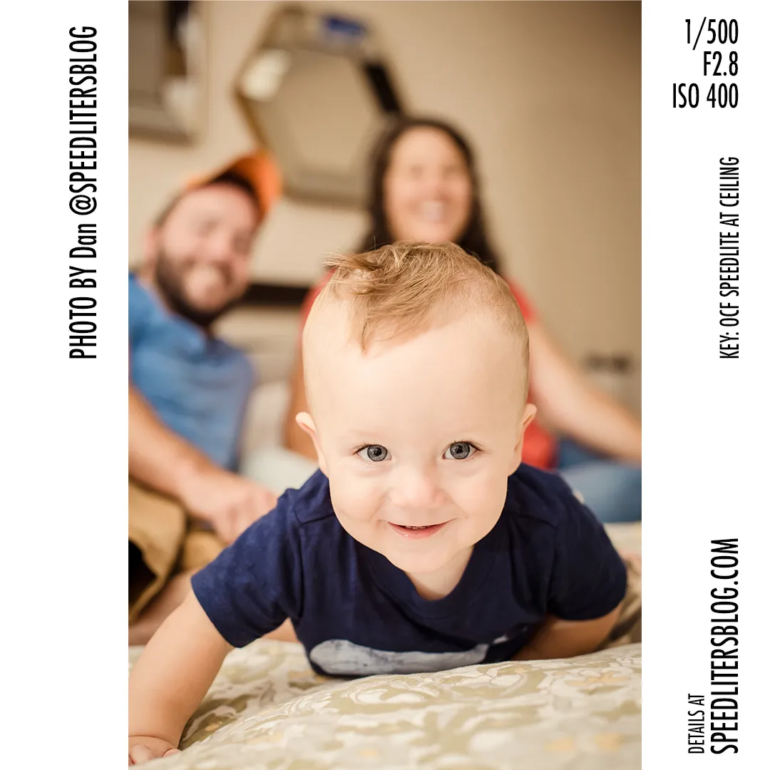Smiling one-year-old crawling on a bed with his parents behind him, lit by a speedlite bounced off the ceiling