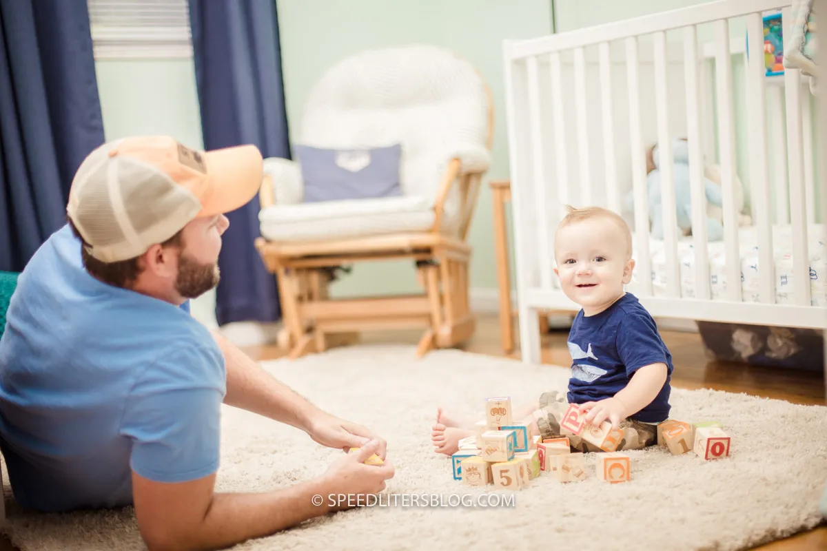 One-year-old playing with alphabet blocks on a nursery rug while his dad watches
