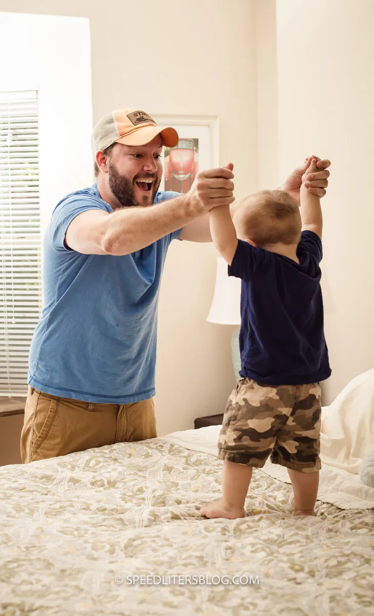 Dad holding his son’s hands as he stands on a bed, both laughing