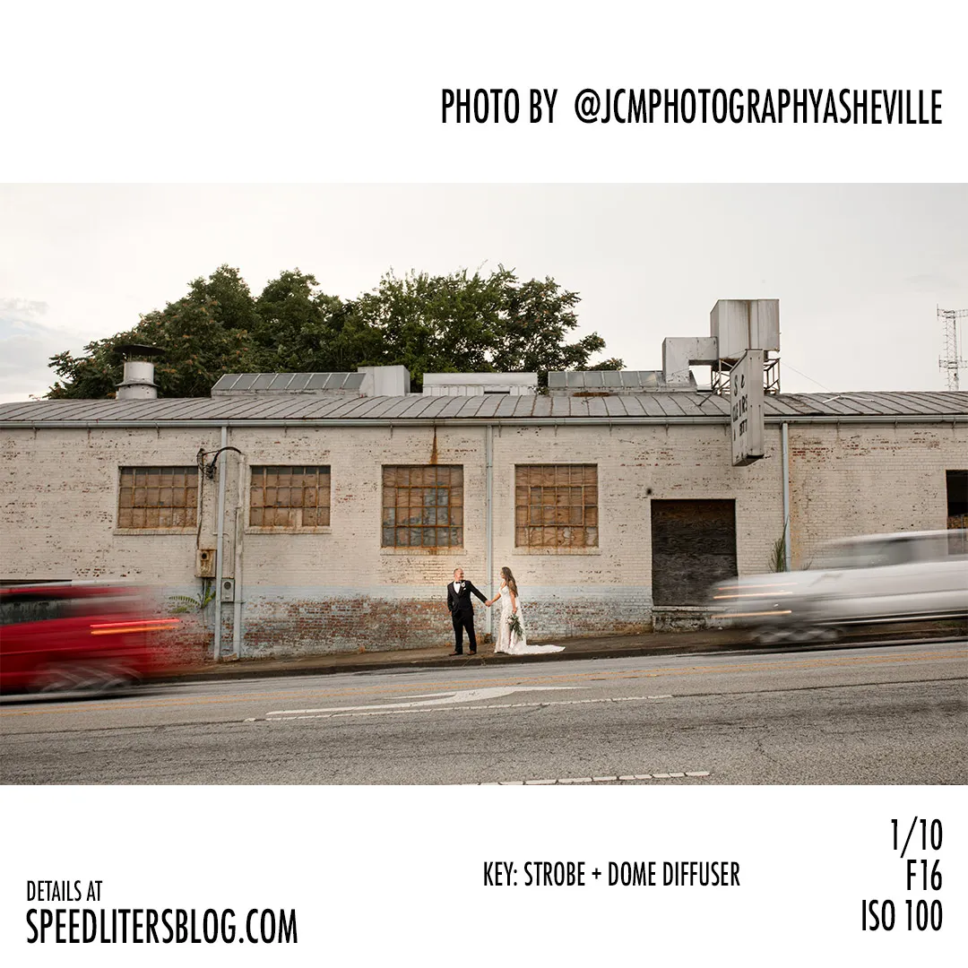 Bride and groom by an old industrial building as cars blur past, by JCM Photography