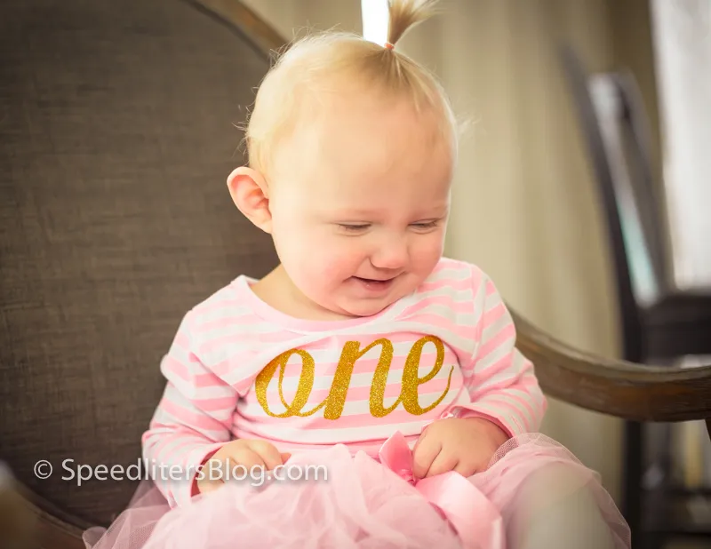 Smiling one-year-old girl in a pink ‘one’ shirt sitting in a chair