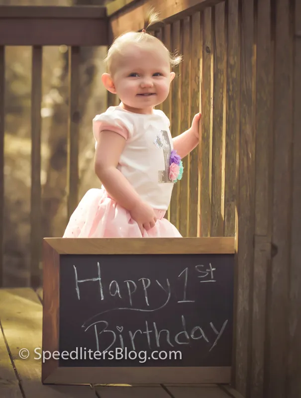 One-year-old standing behind a Happy 1st Birthday chalkboard on a deck