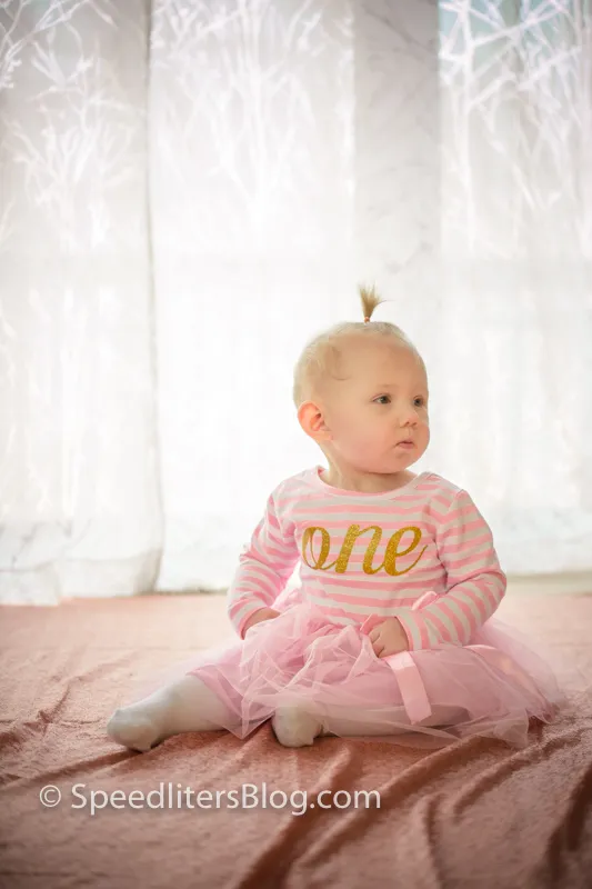 One-year-old girl in a pink tutu sitting by a sheer-curtained window