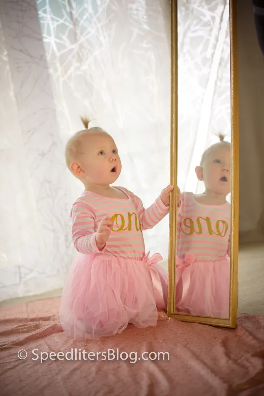 One-year-old girl in a tutu looking at her reflection in a floor mirror