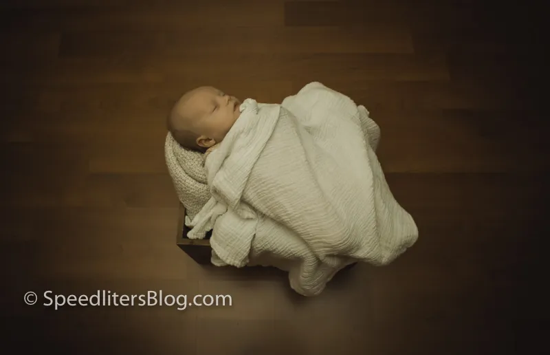 Newborn swaddled in a white blanket on a dark wood floor, shot from above