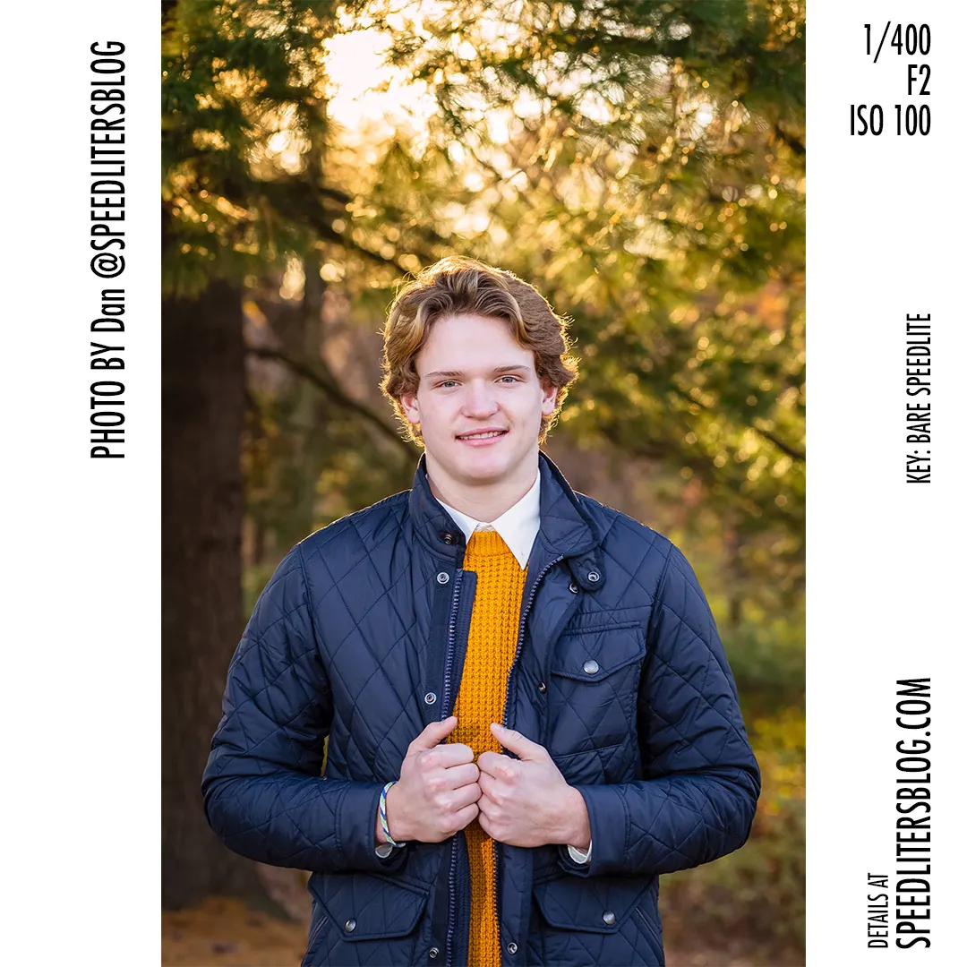 High school senior in a quilted jacket smiling in a fall forest, lit by a bare speedlite