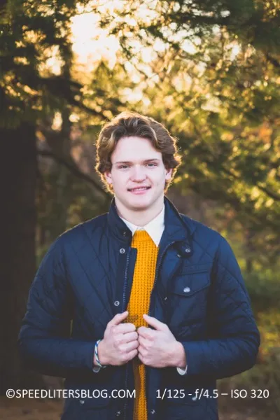 Senior in a quilted jacket and mustard sweater in a fall forest, lit by a bare speedlite
