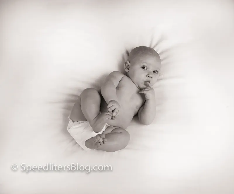 Black-and-white photo of a baby lying on white sheets with a hand to his mouth