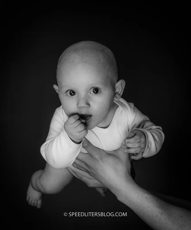 Black-and-white photo of a baby held up against a dark background, chewing his hand